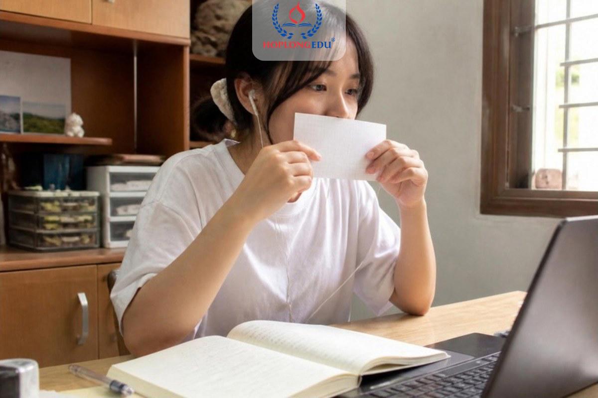 Bạn nữ luyện phát âm tiếng Hàn bên bàn học, tay cầm tờ giấy trước miệng để kiểm tra âm bật hơi