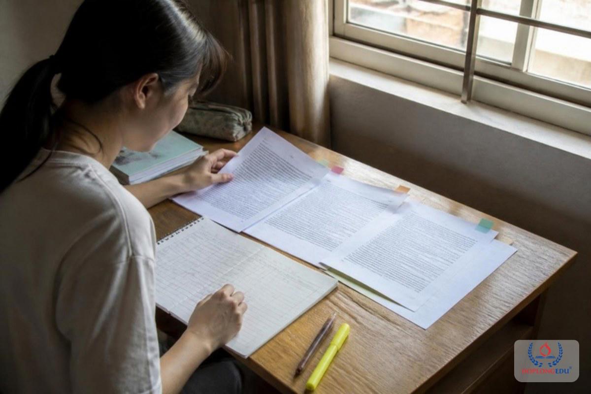Nữ sinh đối chiếu nhiều tờ giấy tờ trên bàn học bằng một bảng ghi tay