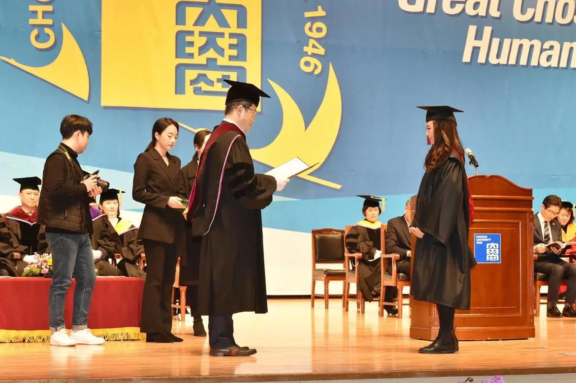 Lễ trao bằng tốt nghiệp tại hội trường Đại học Chosun, phía sau là biểu tượng trường và năm thành lập 1946