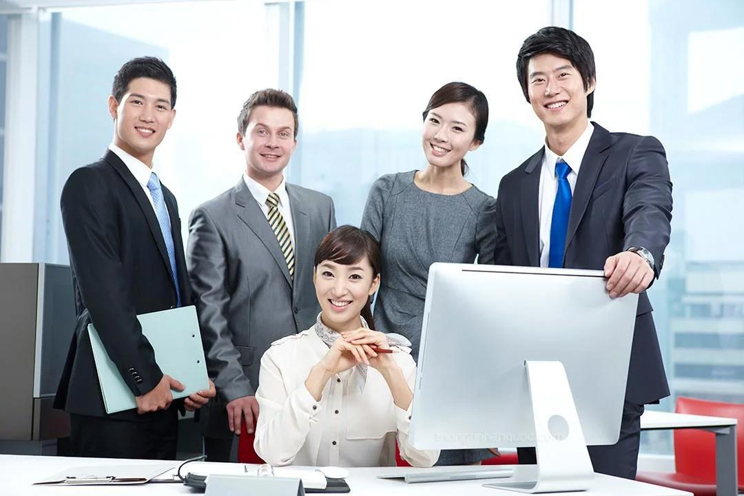 Nhóm nhân viên văn phòng mặc vest đứng quanh bàn làm việc có máy tính